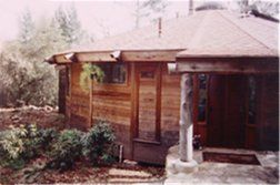 Wooden cabin with a porch, shingled roof, and a stone chimney amid trees and shrubs.