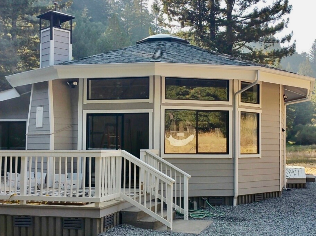 Small beige round house with white porch railing and deck in a wooded setting, labeled