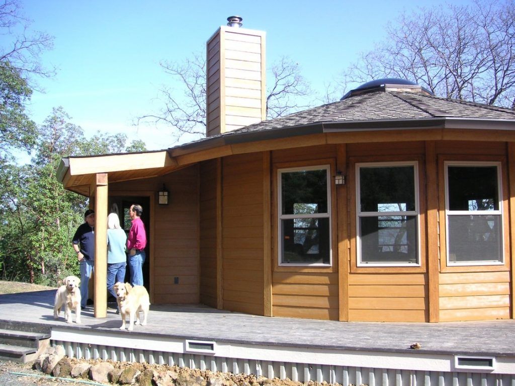 People standing outside a small wooden building with large windows and a stone chimney.