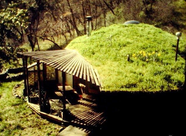 Rustic wooden shelter beside a grassy mound in a wooded park, with a bench and path