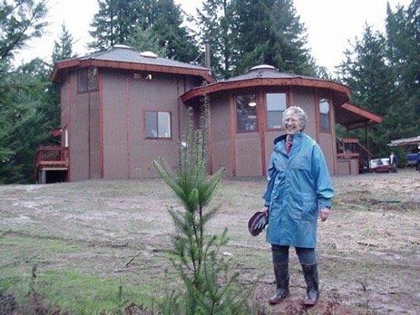 Person in a blue raincoat stands outside a rustic cabin among trees.