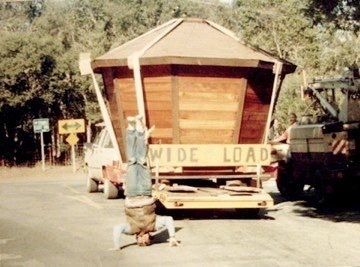 Wide-load house on a trailer being transported down a road, with people nearby.