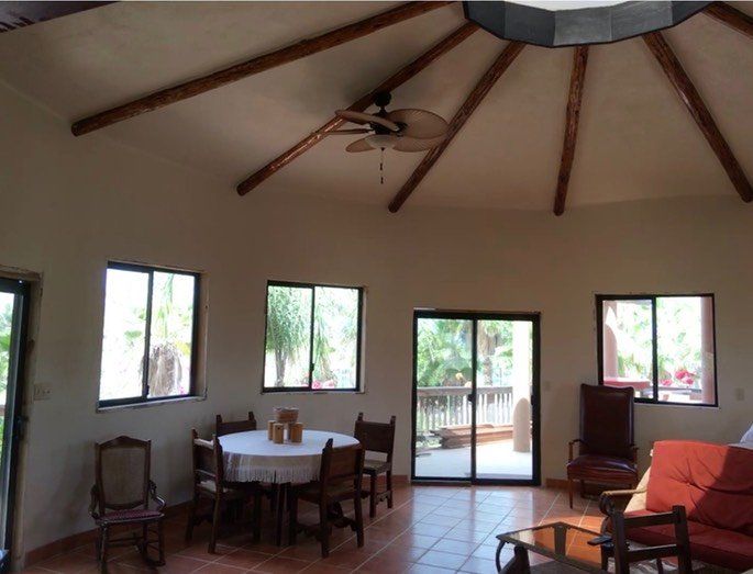 Bright living room with wooden ceiling beams, a dining table, chairs, windows, and a red sofa.