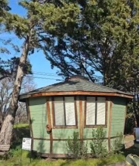 Small round green building with brown trim and a shingled roof among trees outdoors
