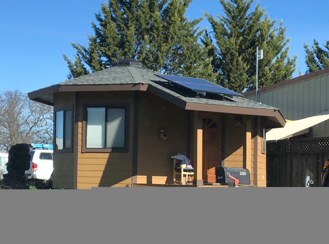 Small brown house with solar panels, blue sky, and tall trees in the background