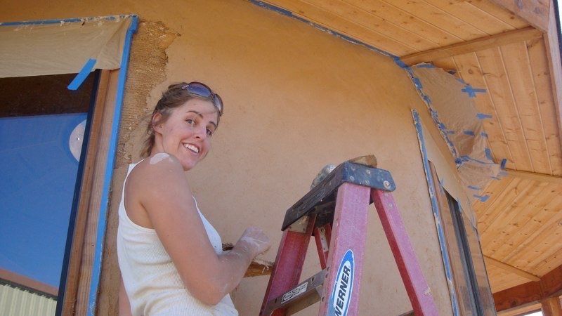Person on a ladder painting an orange wall, smiling toward the camera.