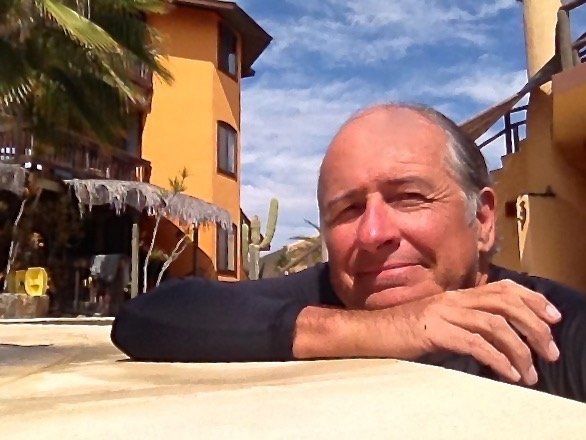 Man leaning on a pool edge in a sunny courtyard with orange buildings and palm trees nearby