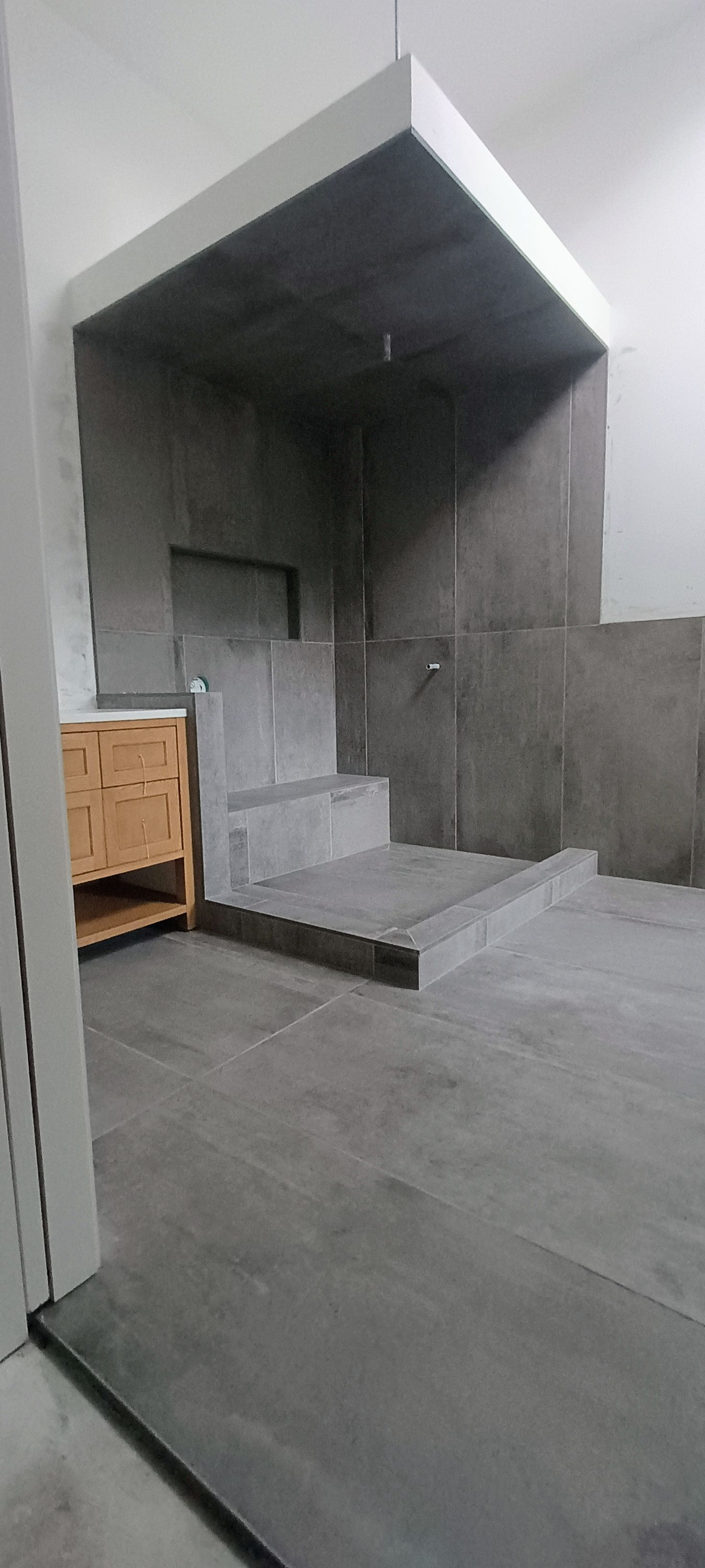 A modern walk-in shower with a built-in stone bench, featuring dark gray tile walls and floor, and a light wood cabinet.