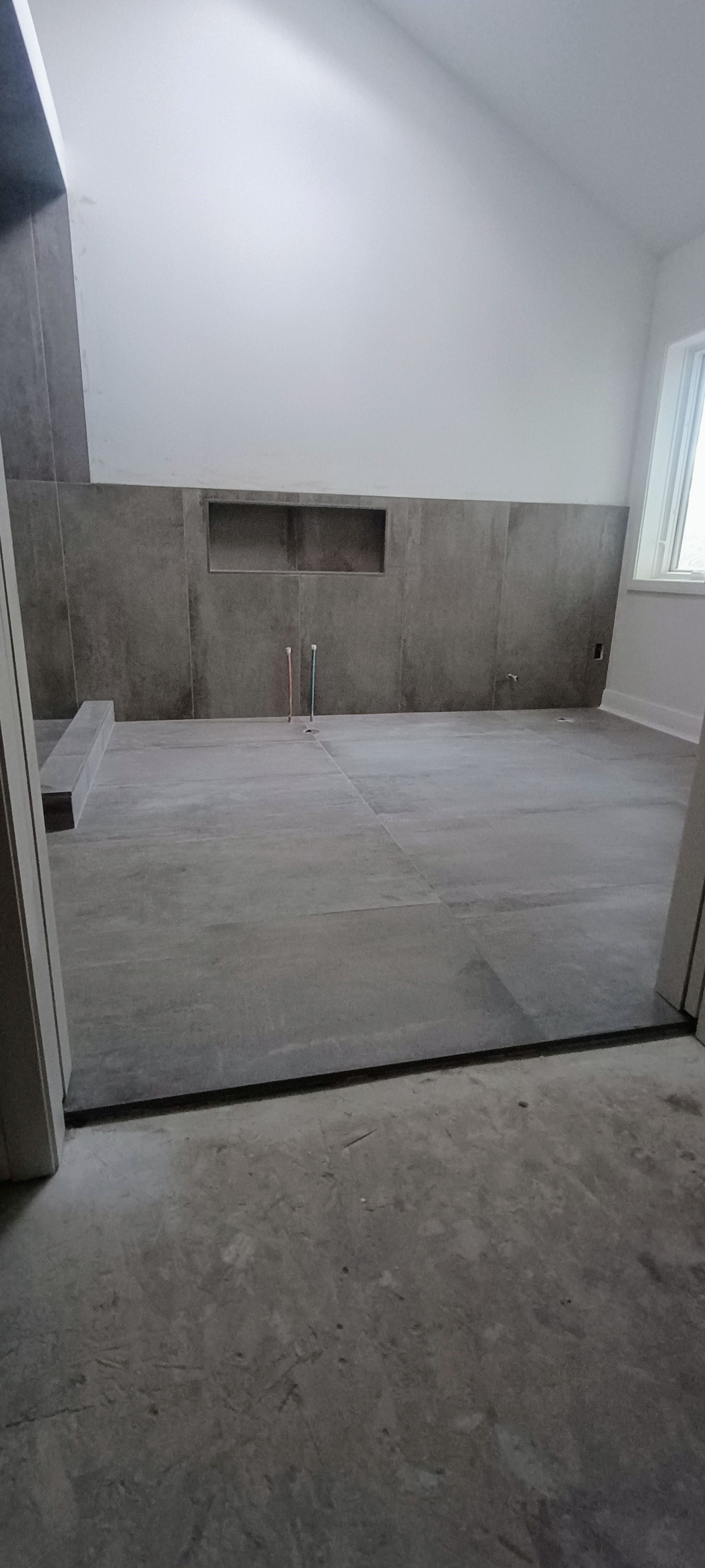 An unfinished room with large grey floor and wall tiles, a recessed niche in the back wall, and exposed plumbing pipes.