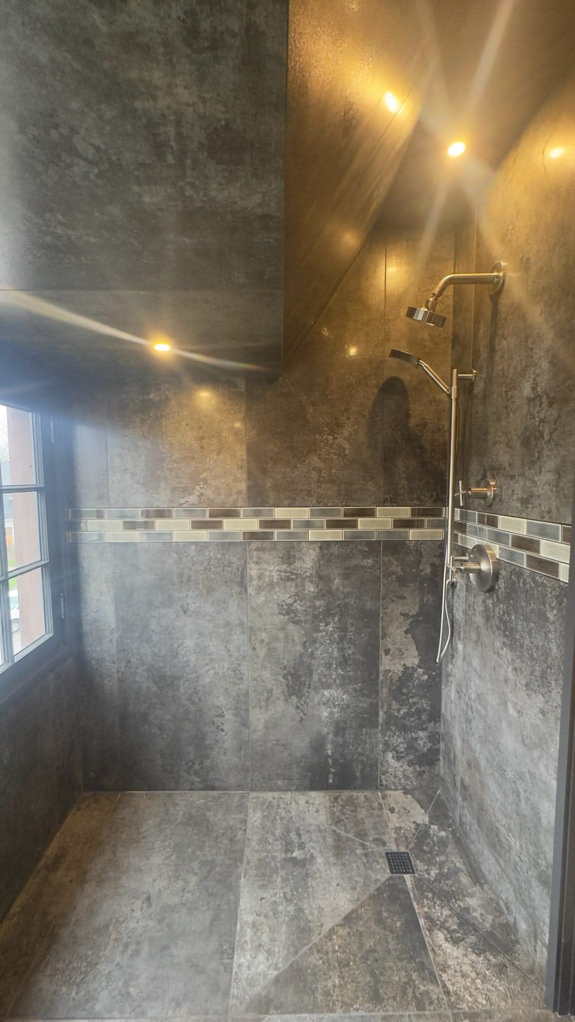 A modern walk-in shower with dark textured tile walls, a decorative mosaic stripe, and a brushed nickel shower head.