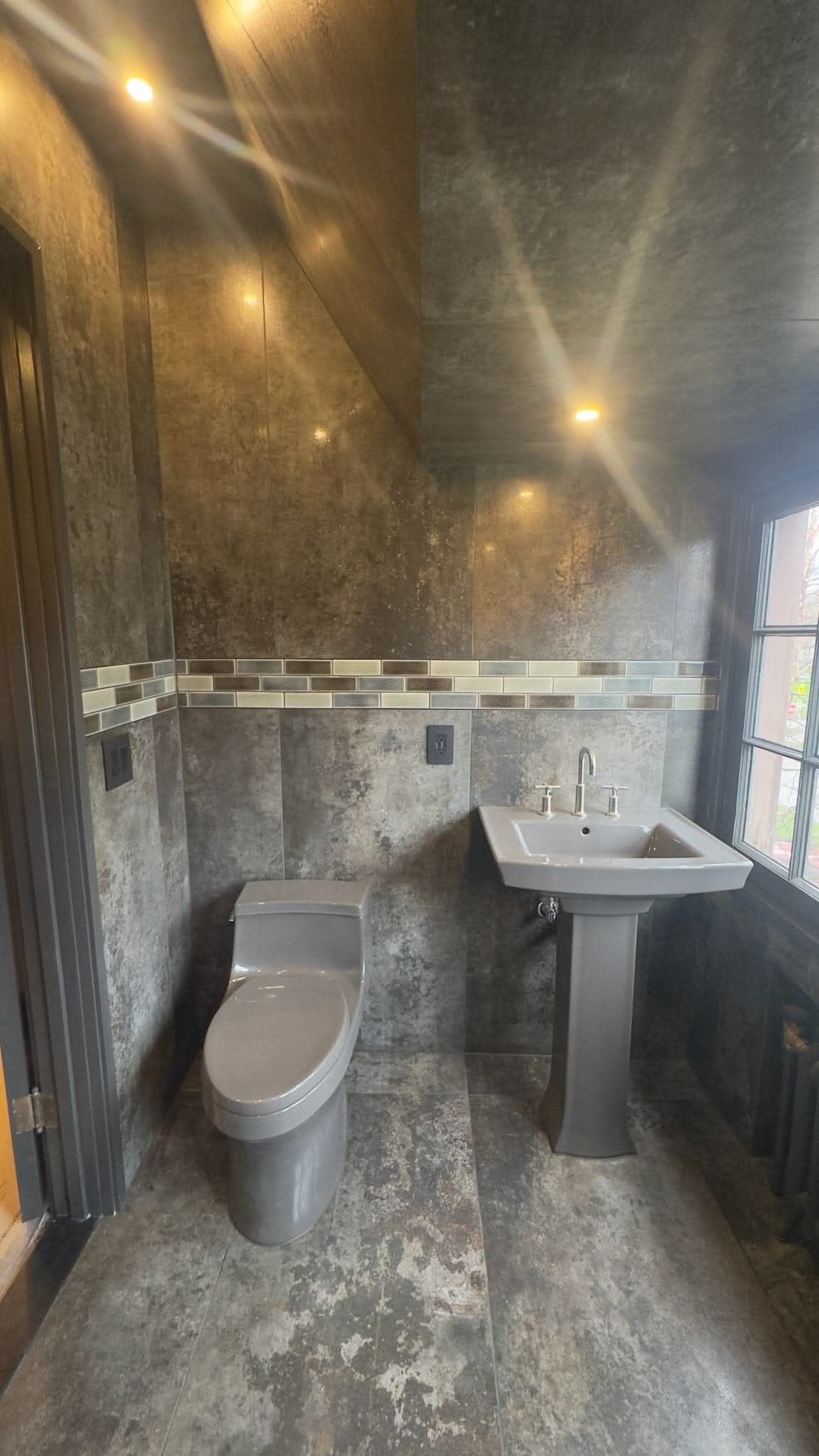 Another side of modern walk-in shower with dark textured tile walls, a decorative mosaic stripe, and a grey toilet and sink.
