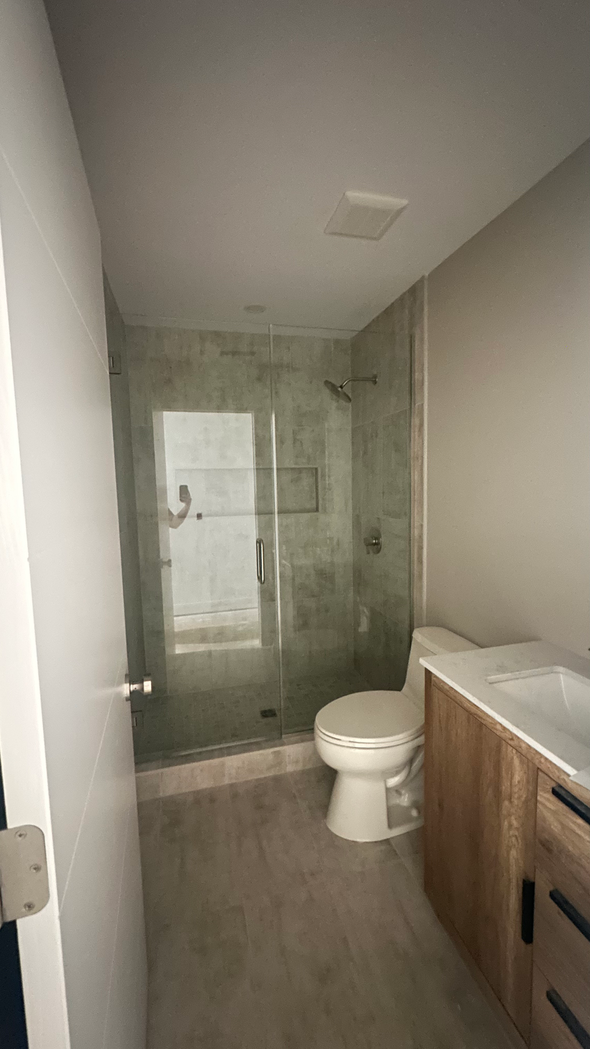 A modern bathroom with a light wood vanity, a toilet, and a glass-enclosed shower with gray tiled walls.