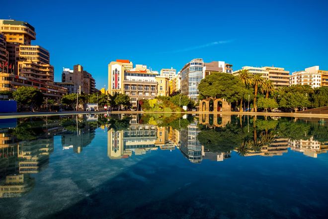 Waterfront city skyline with modern buildings reflected in calm blue water under a clear sky