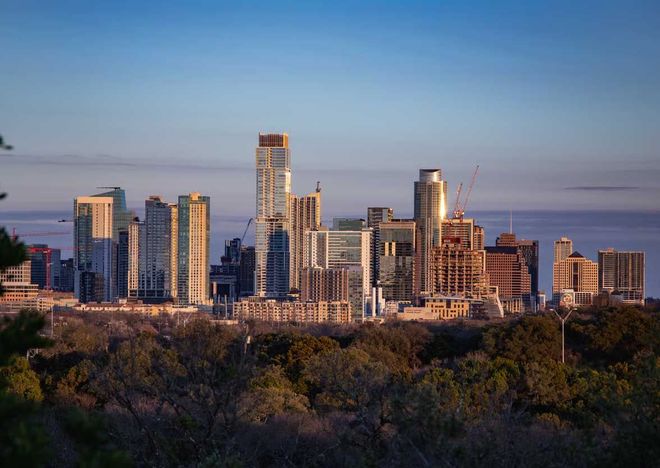 City skyline at sunset with tall modern buildings above a tree-lined foreground