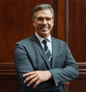Smiling man in a gray suit with crossed arms, standing against a wooden-paneled wall.