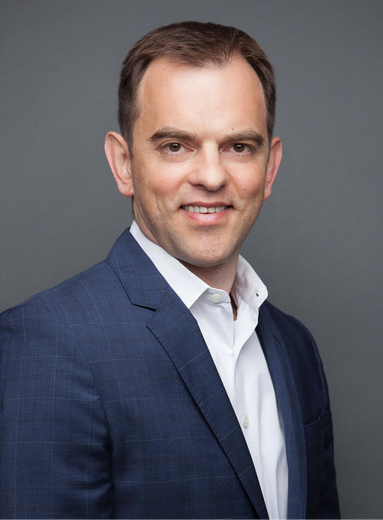 Professional headshot of a person in a blue suit and white shirt against a gray background