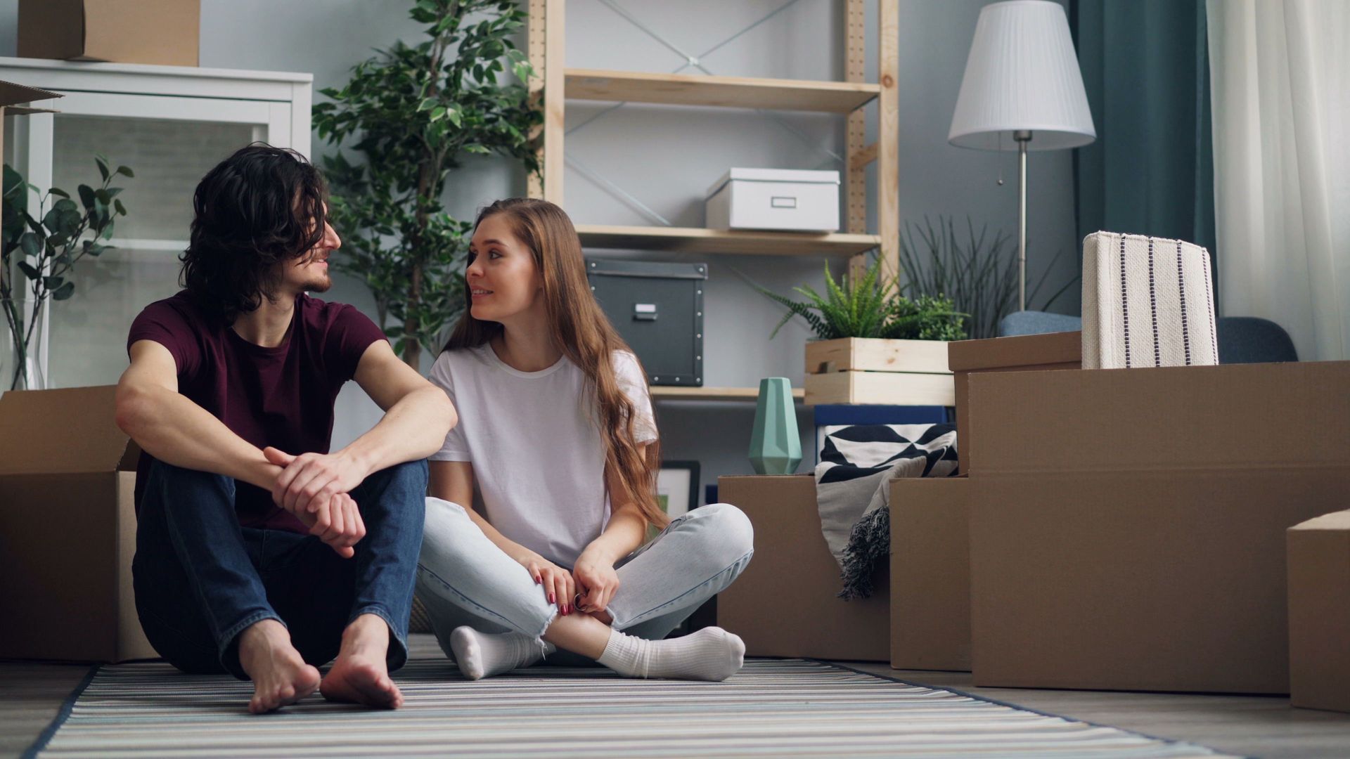 Two people sitting on the floor in a living room among moving boxes, talking and smiling.