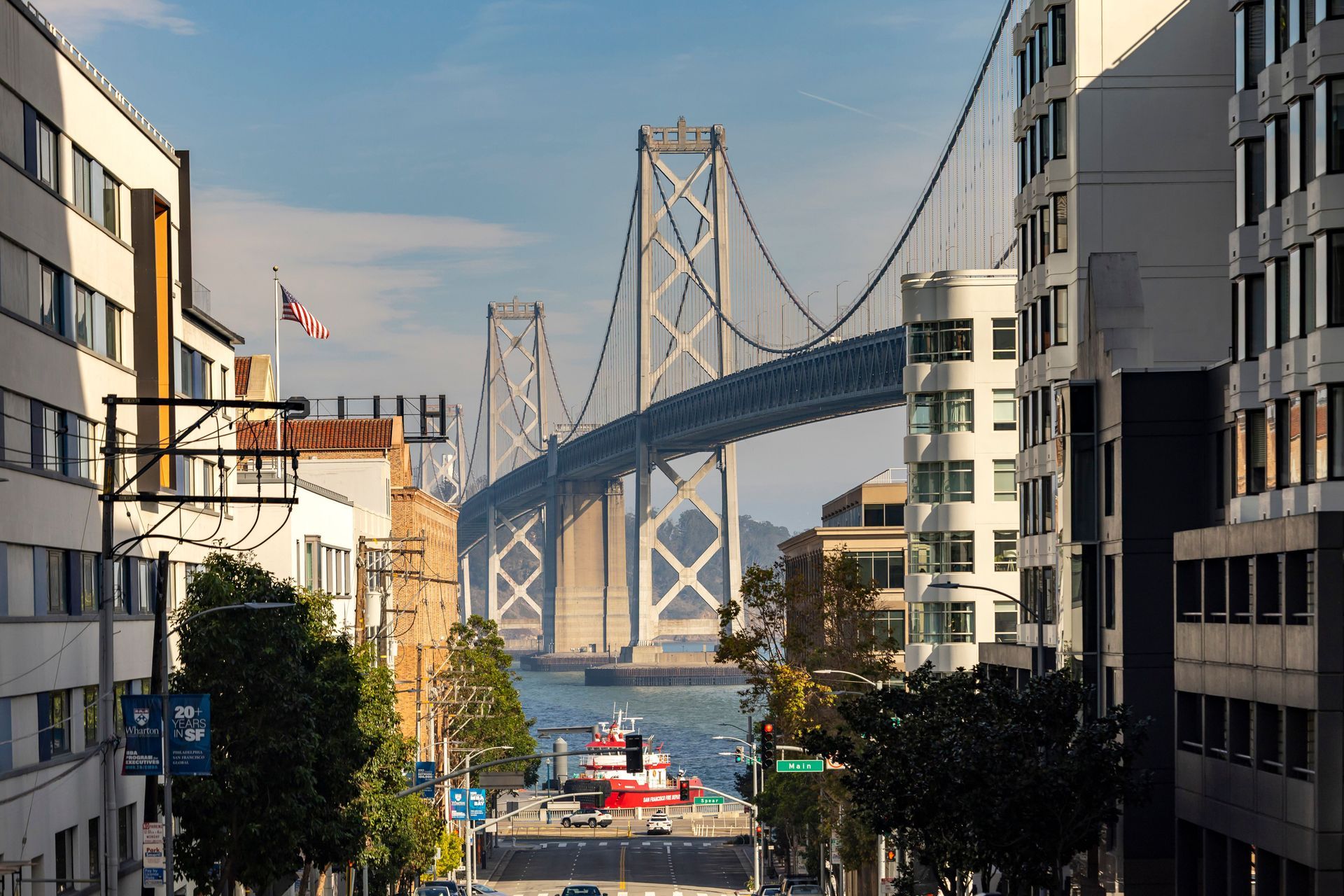 Suspension bridge over a city street between buildings, with water and boats below in sunlight