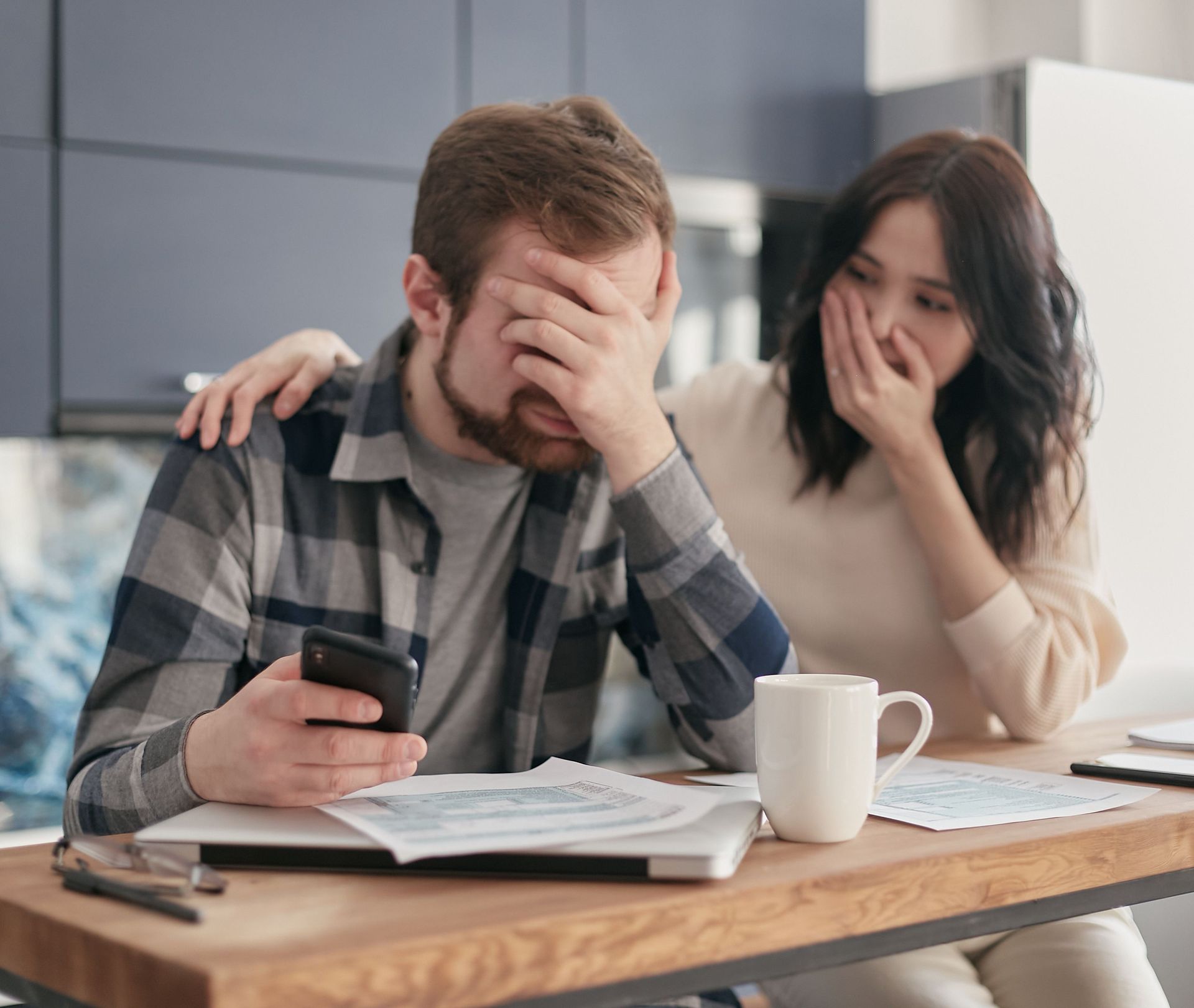 Two people at a table, both covering their faces with worry while looking at papers and a phone.