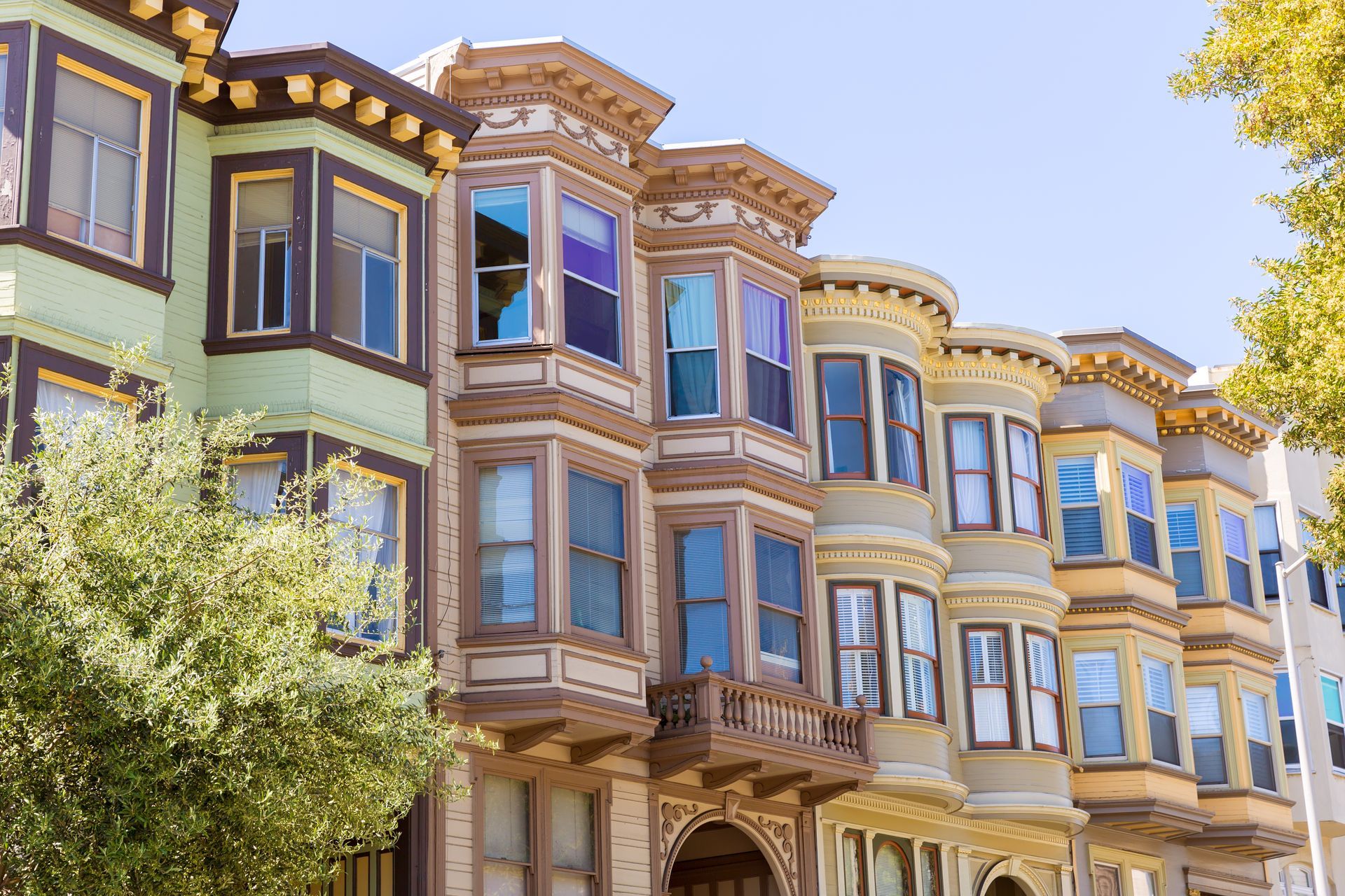 Colorful Victorian-style row houses with bay windows under a clear blue sky, framed by leafy trees