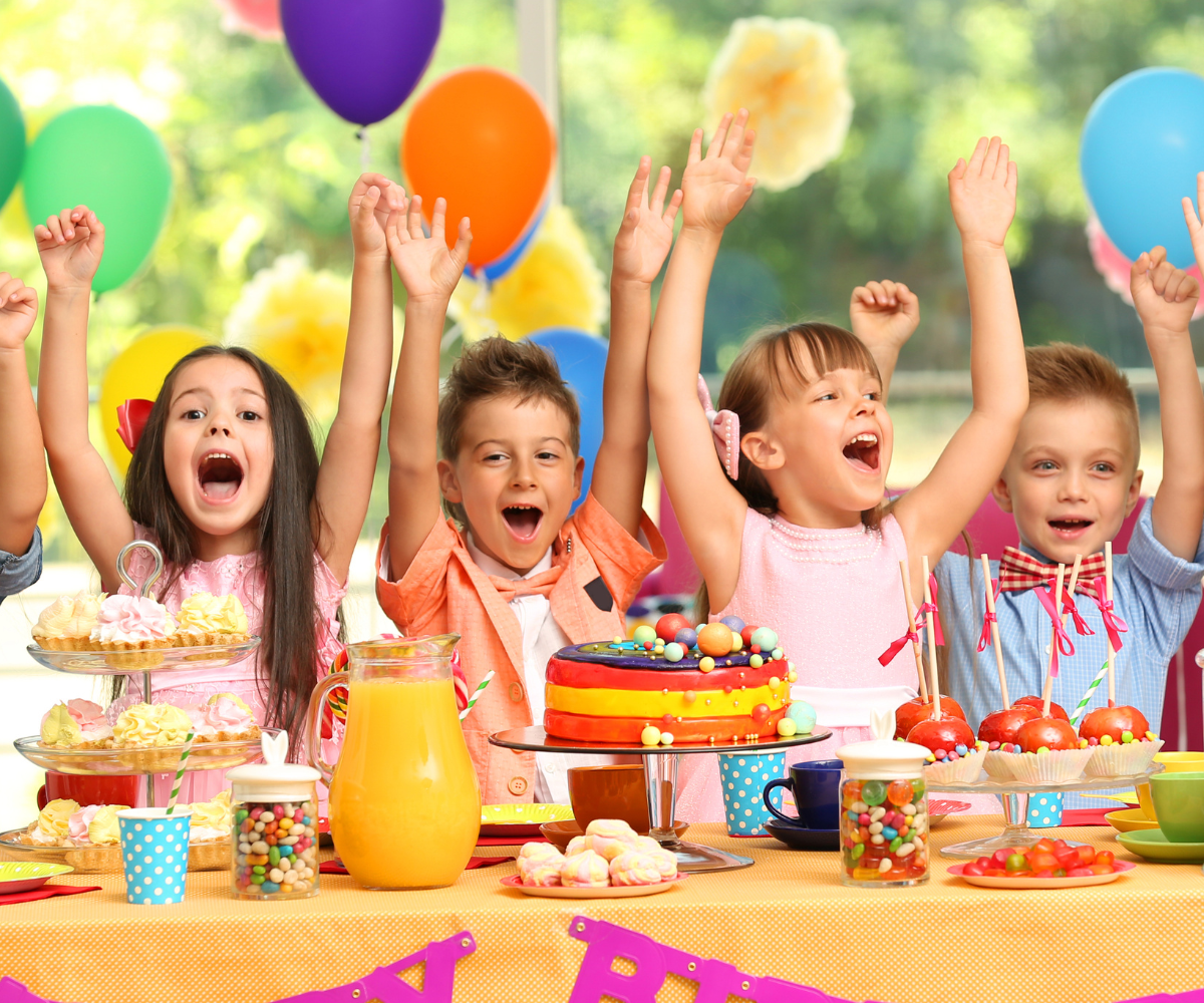 A group of children are sitting at a table with their arms in the air at a birthday party.
