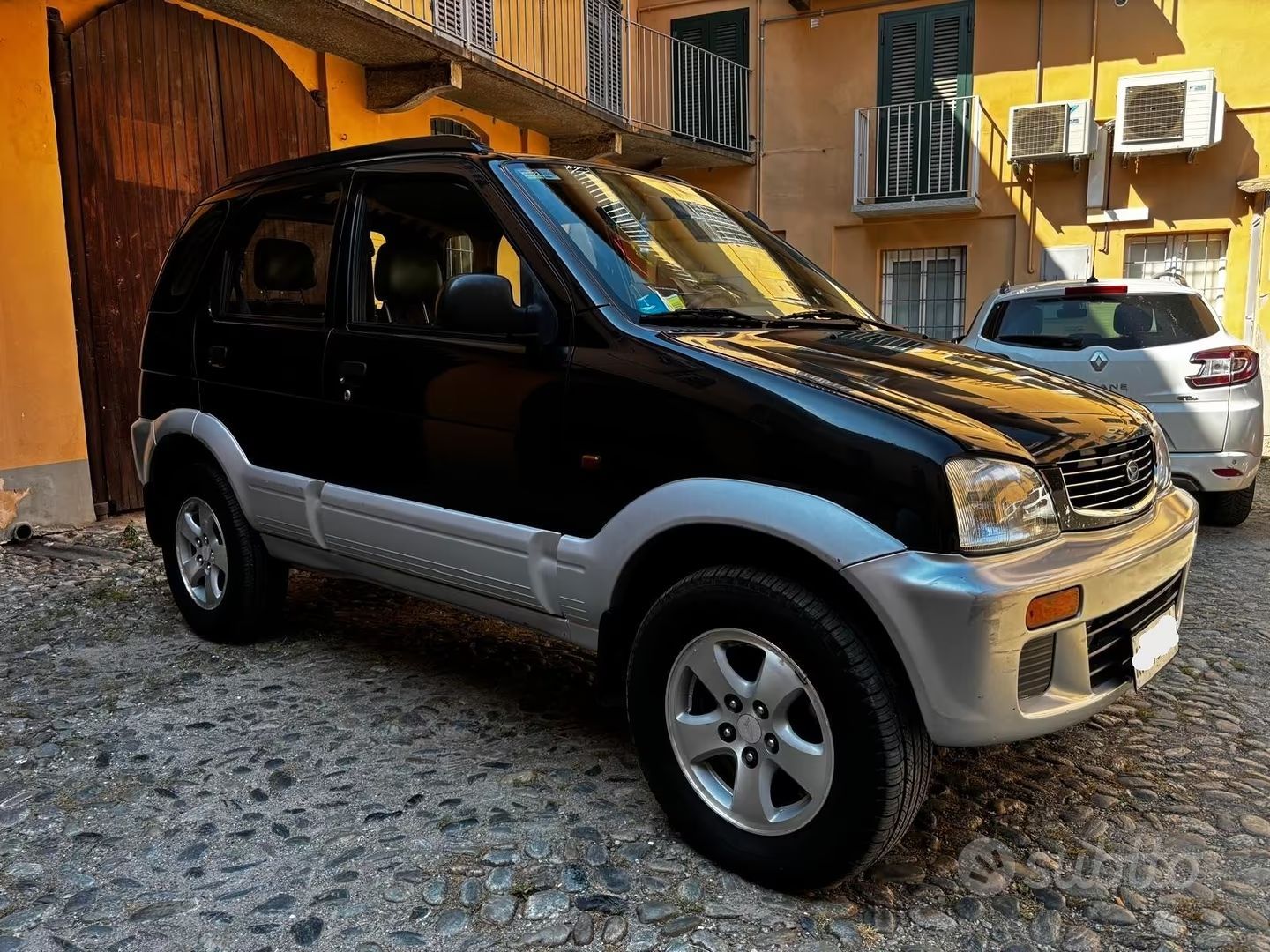 Un SUV argento e nero è parcheggiato su una strada acciottolata accanto a edifici gialli.