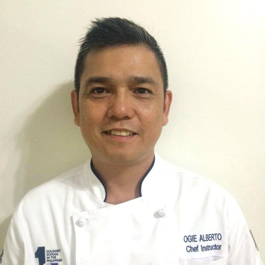 Smiling chef in a white coat against a plain light background.