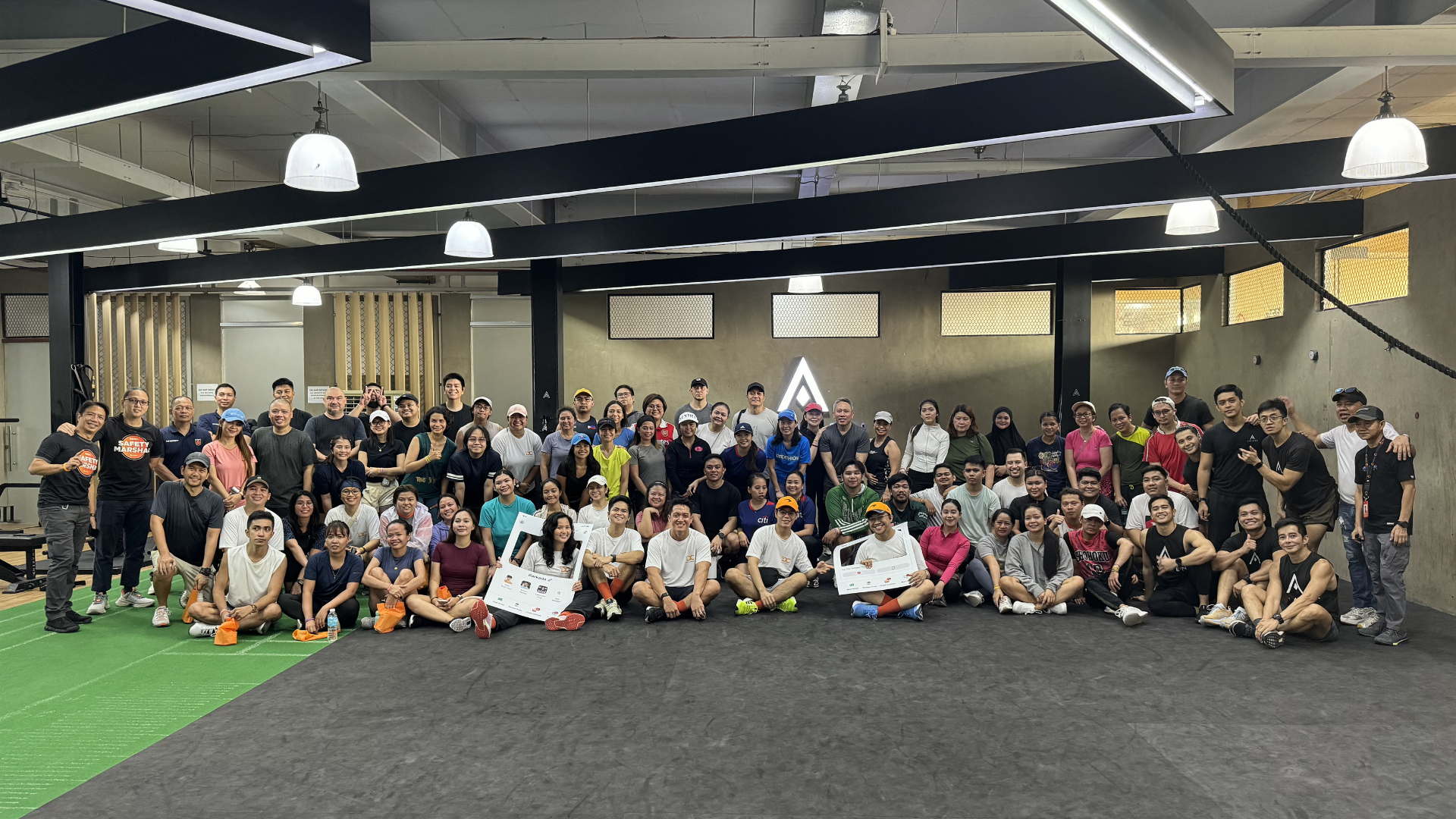 Large group posing in an indoor gym, seated and standing on a dark floor under hanging lights.