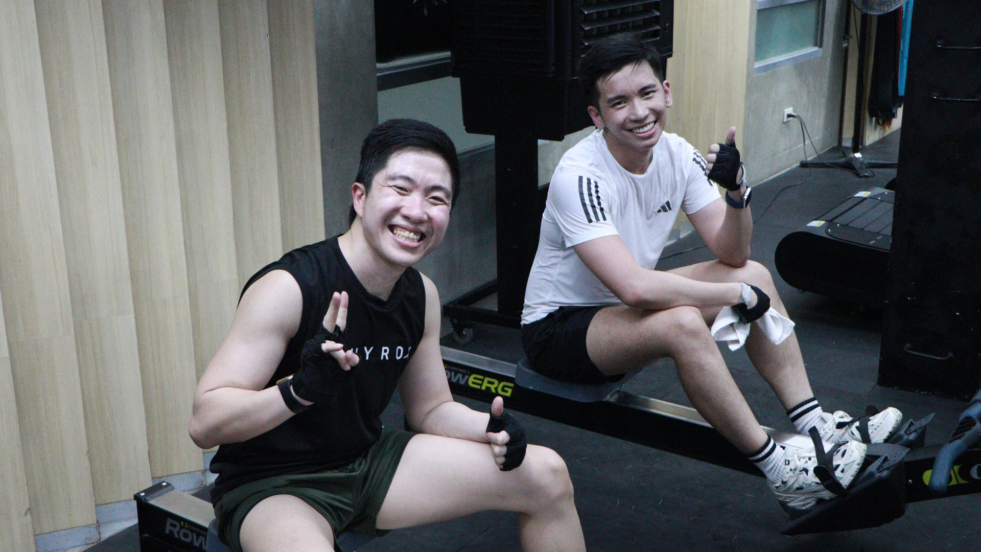 Two smiling people sitting on steps indoors, one giving a thumbs-up and the other holding a drink.