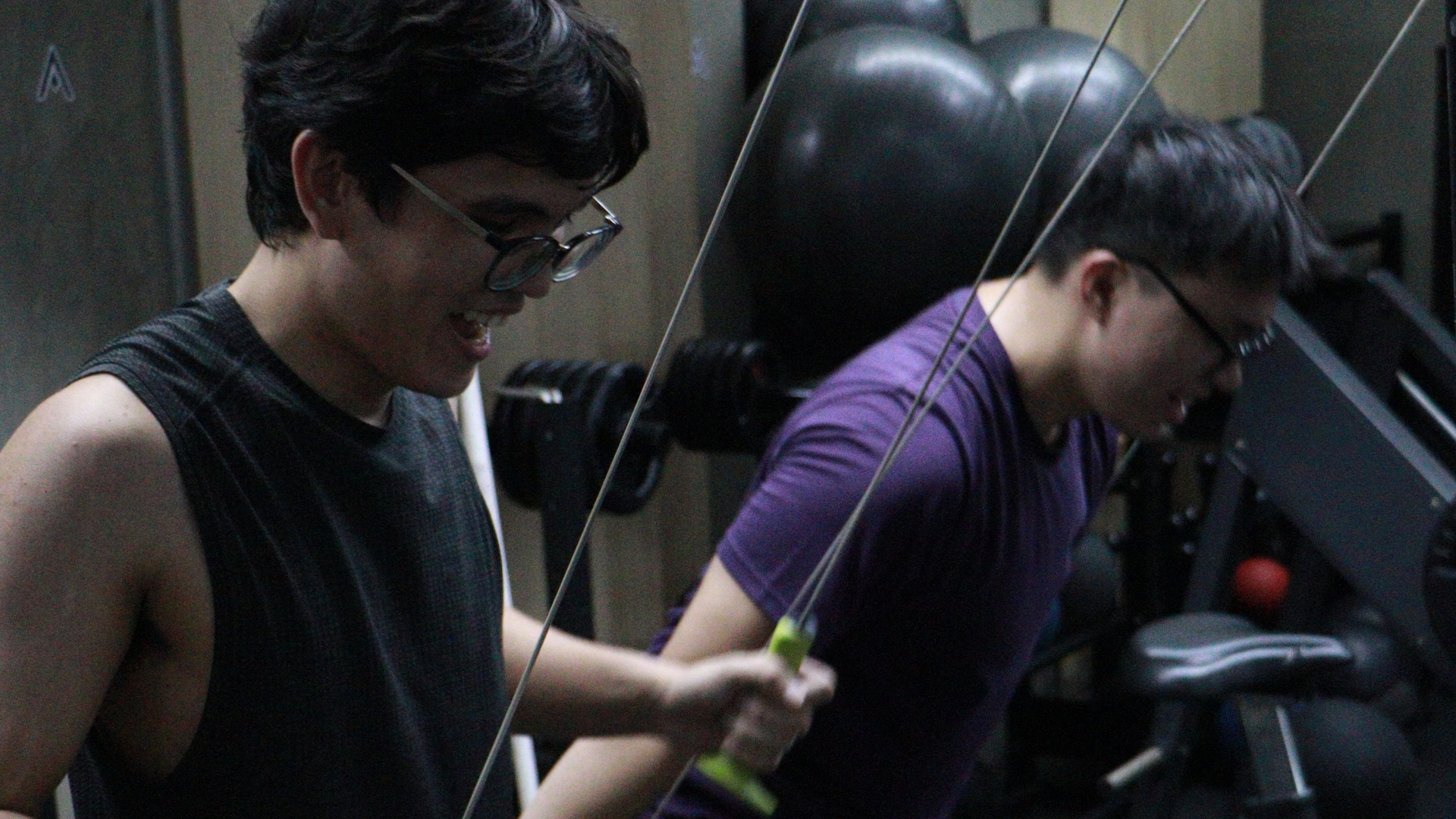 Two people exercising in a gym, one in a black tank top and one in a purple shirt, near a weight machine.