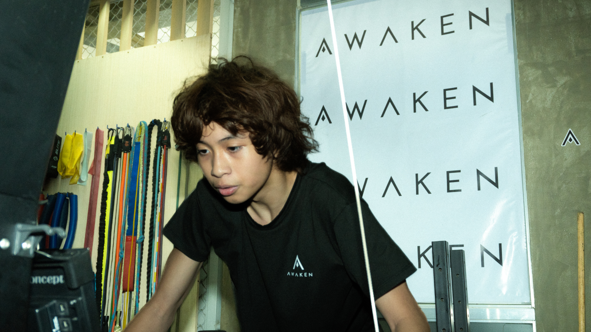 Person leaning forward beside a sign reading “AWAKEN,” with colorful hanging strips in the background