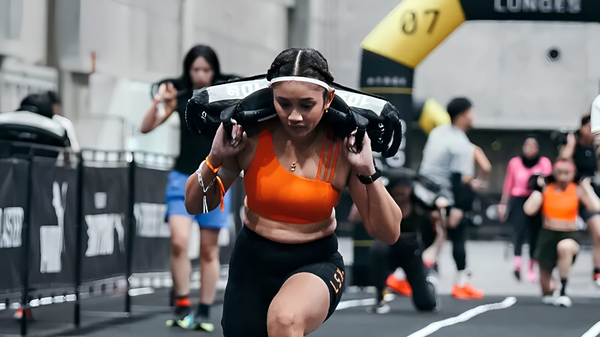 Runner in orange shirt performing sandbag lunges toward the finish line on a HYROX race course
