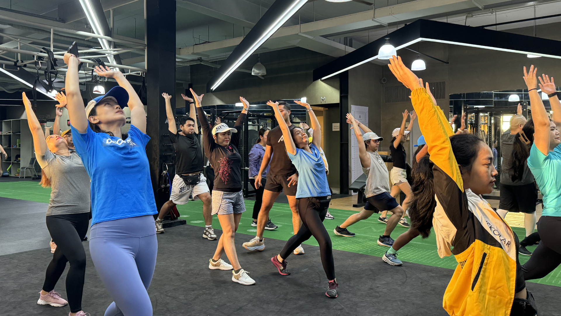 Group fitness class doing overhead stretches in a modern gym, with people in colorful workout clothes.