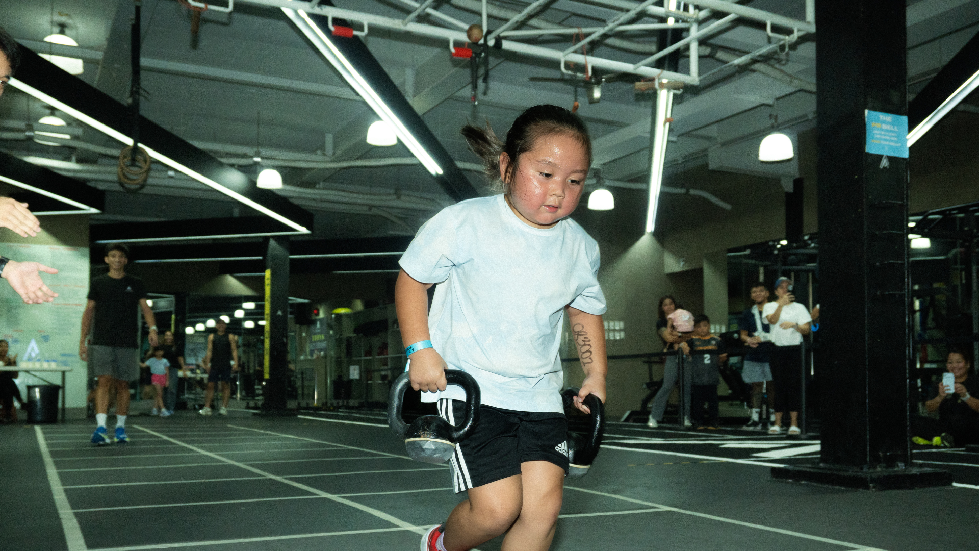 Runner in a gym mid-stride, wearing a white shirt and black shorts, with a race lane and spectators in the background.