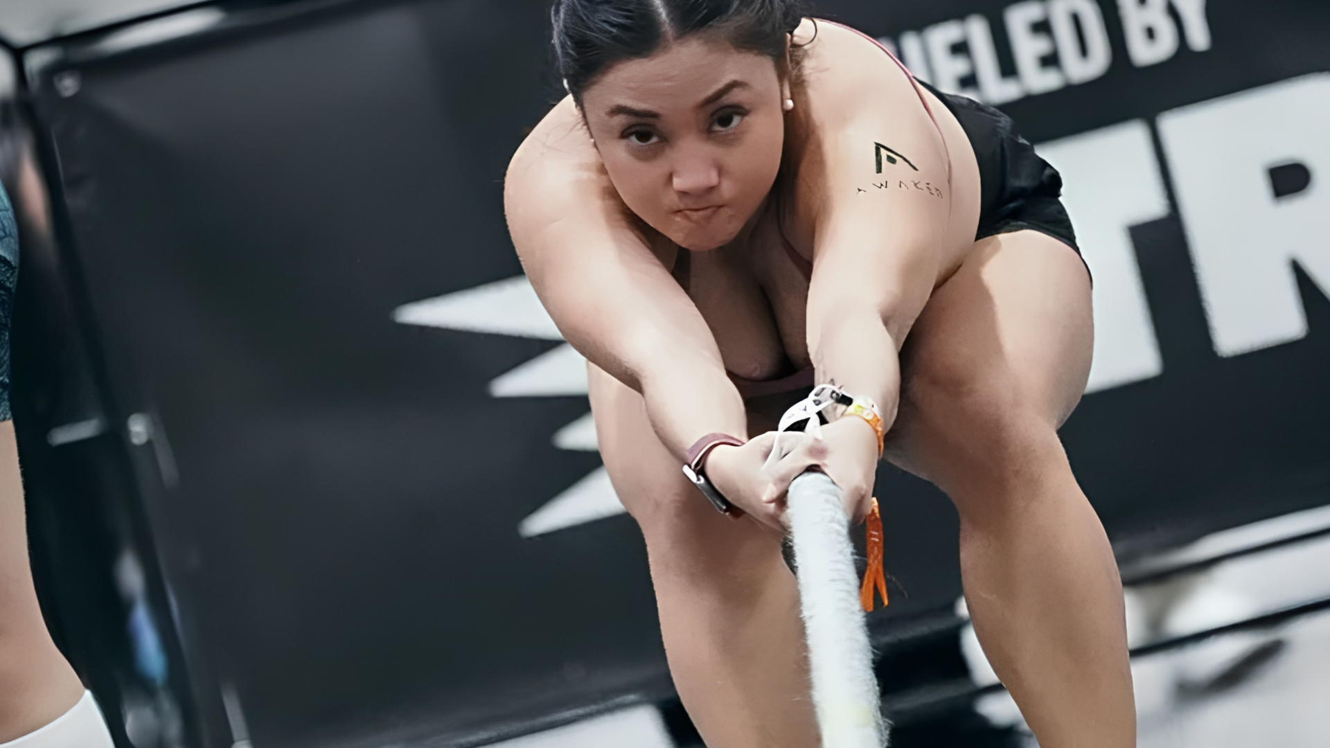 Athlete pulling a thick rope in a competitive HYROX event, crouched low on a black floor.
