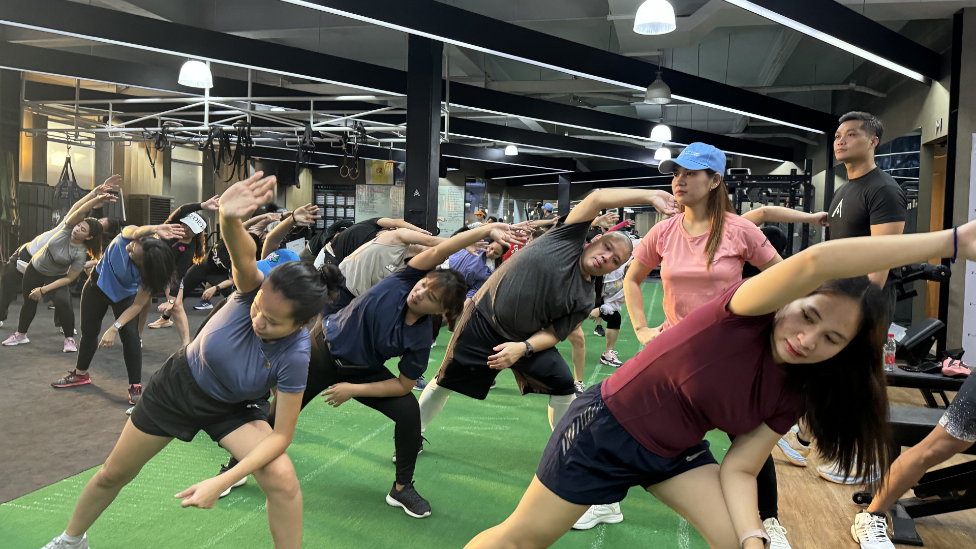People stretching in a gym class on green turf under bright lights