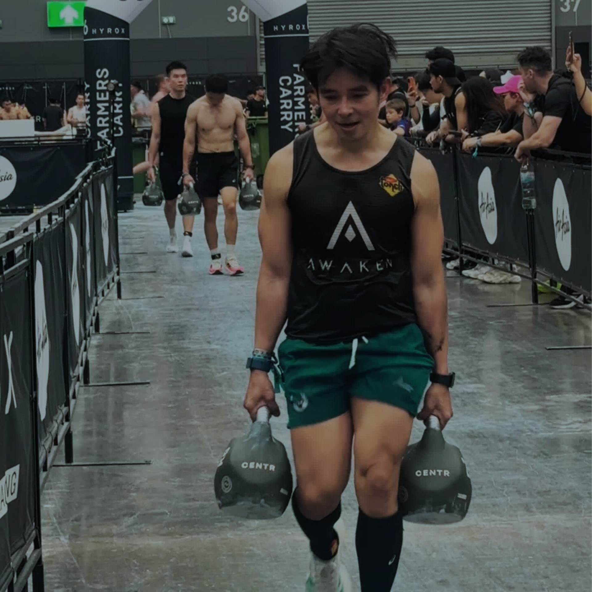 Athlete carrying two kettlebells at a wet indoor obstacle race finish line, with spectators behind barriers