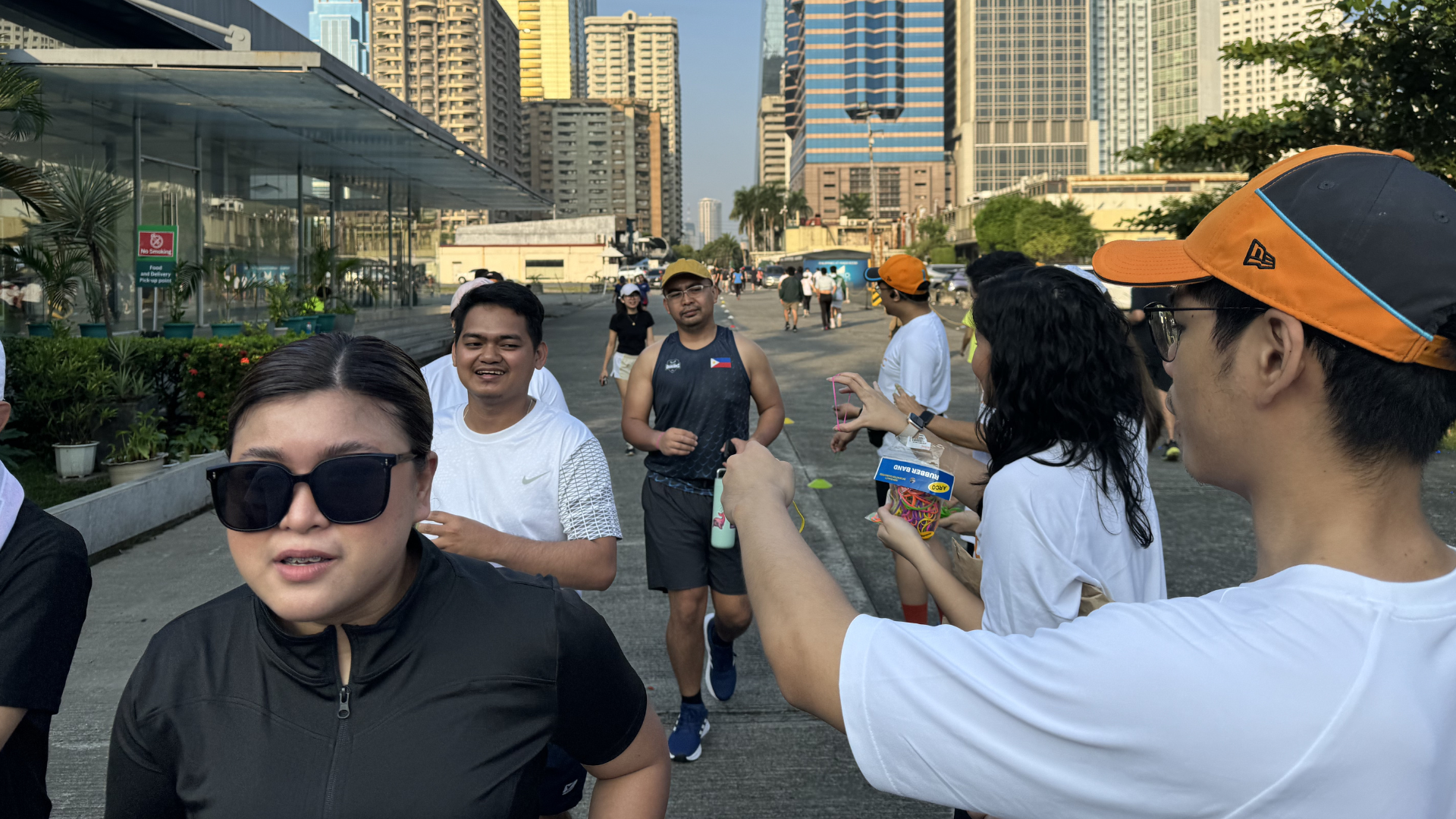 People walking and socializing outdoors in a city plaza, with tall buildings and a waterfront in the background.