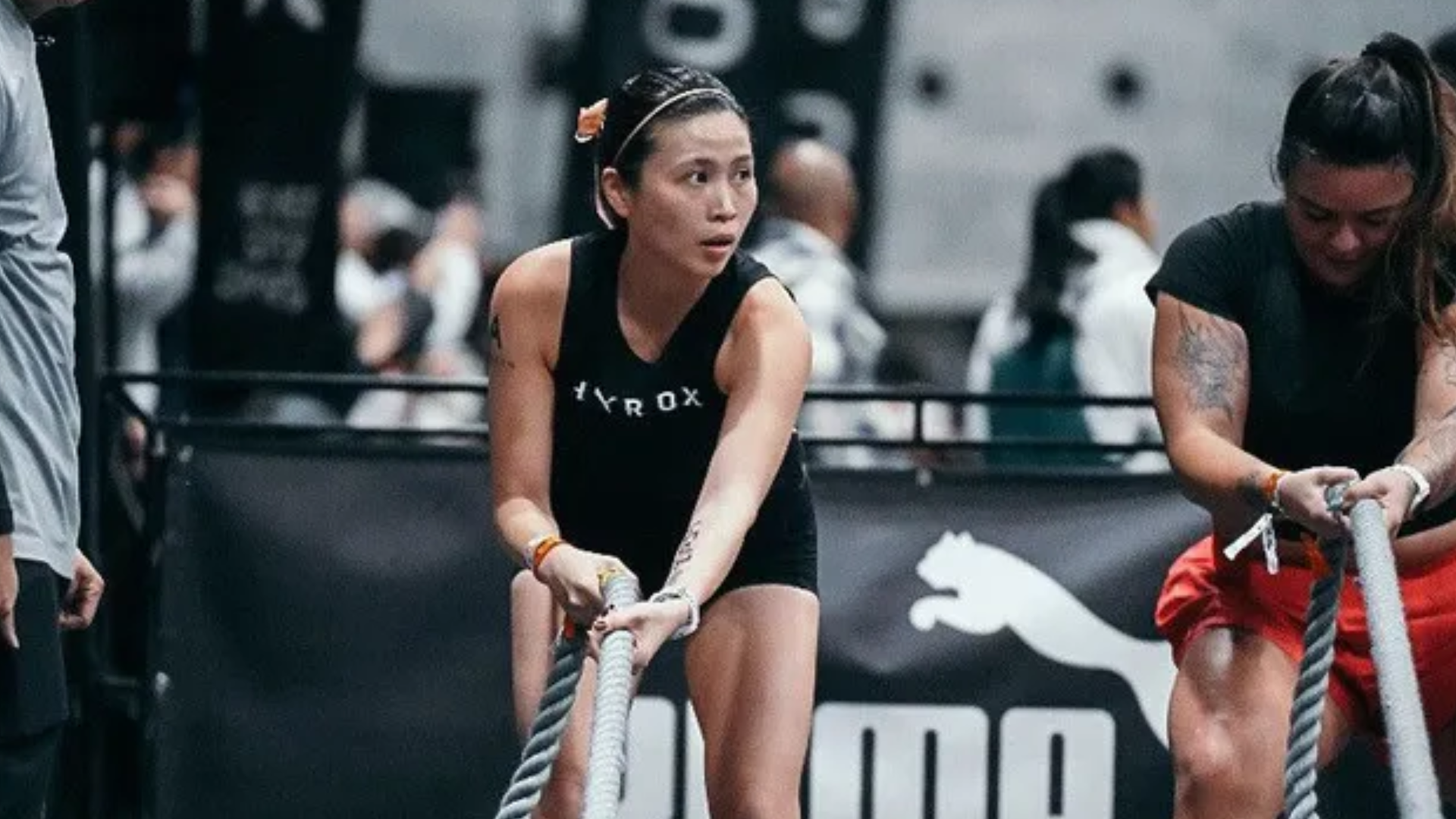 Three athletes in a HYROX competition pulling battle ropes, with a woman centered in black and pink shoes.