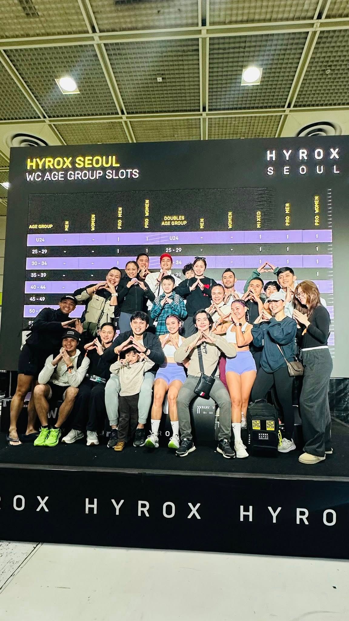 Group posing in front of a HYROX event backdrop with purple lights and a black floor banner.