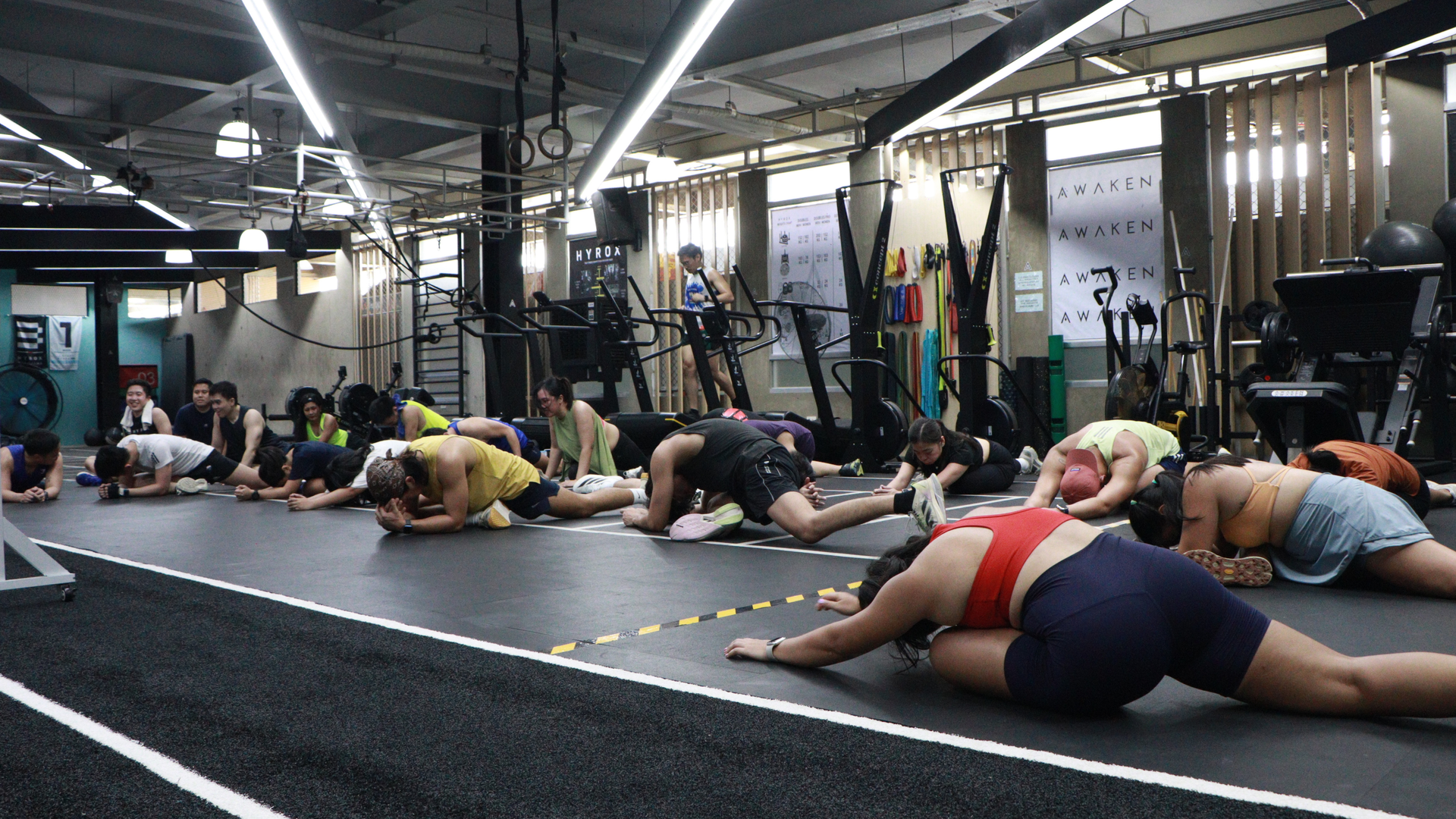People stretching on mats in a gym, with exercise equipment and hanging flags in the background.