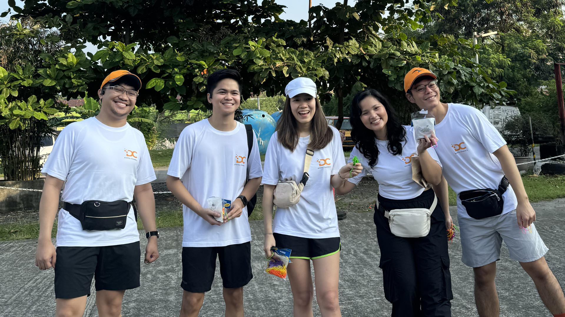 Five friends in white shirts and caps posing outdoors on a sunny path with greenery behind them