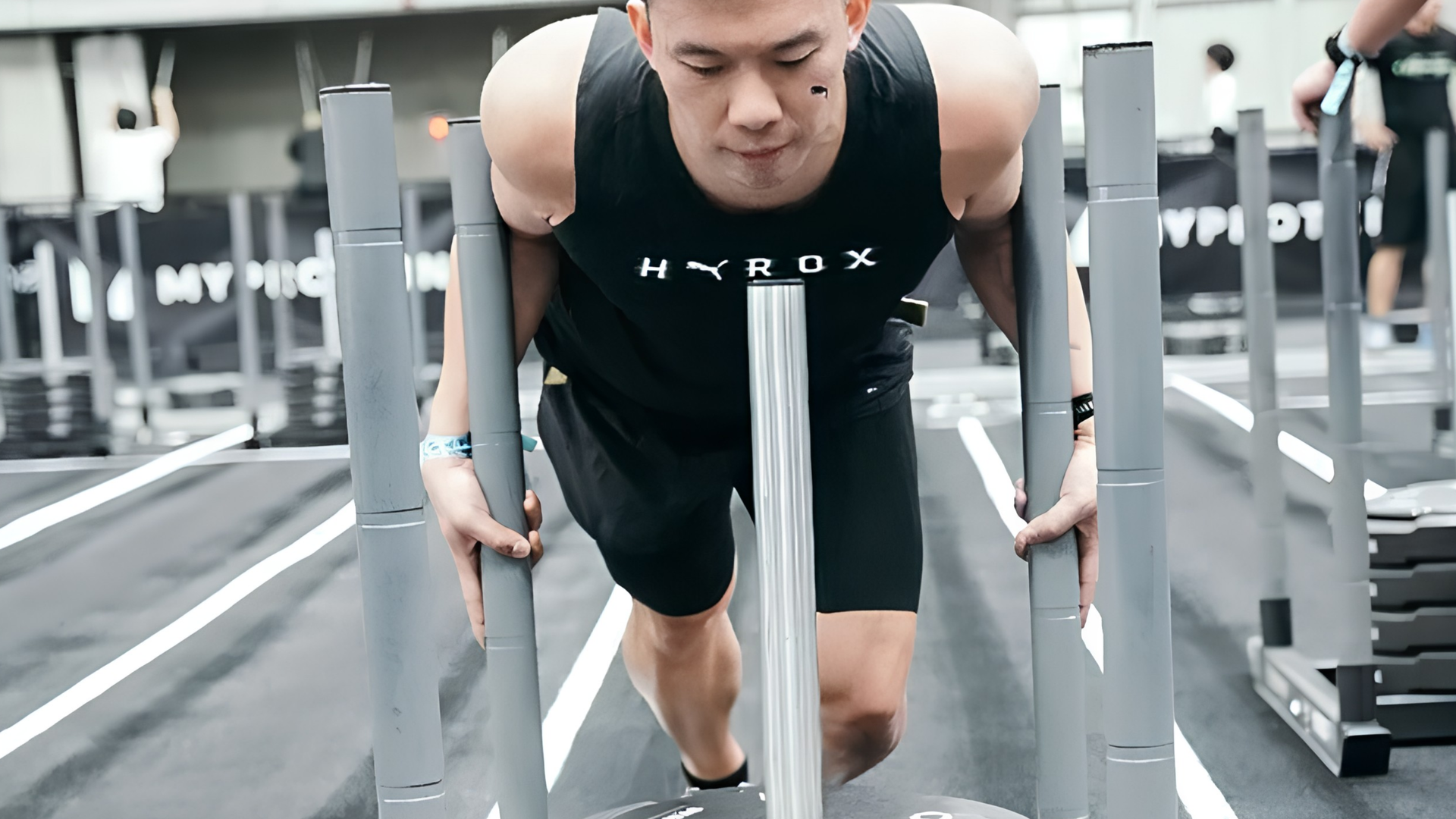 Man pushing a weighted sled in a HYROX competition, leaning forward with effort.