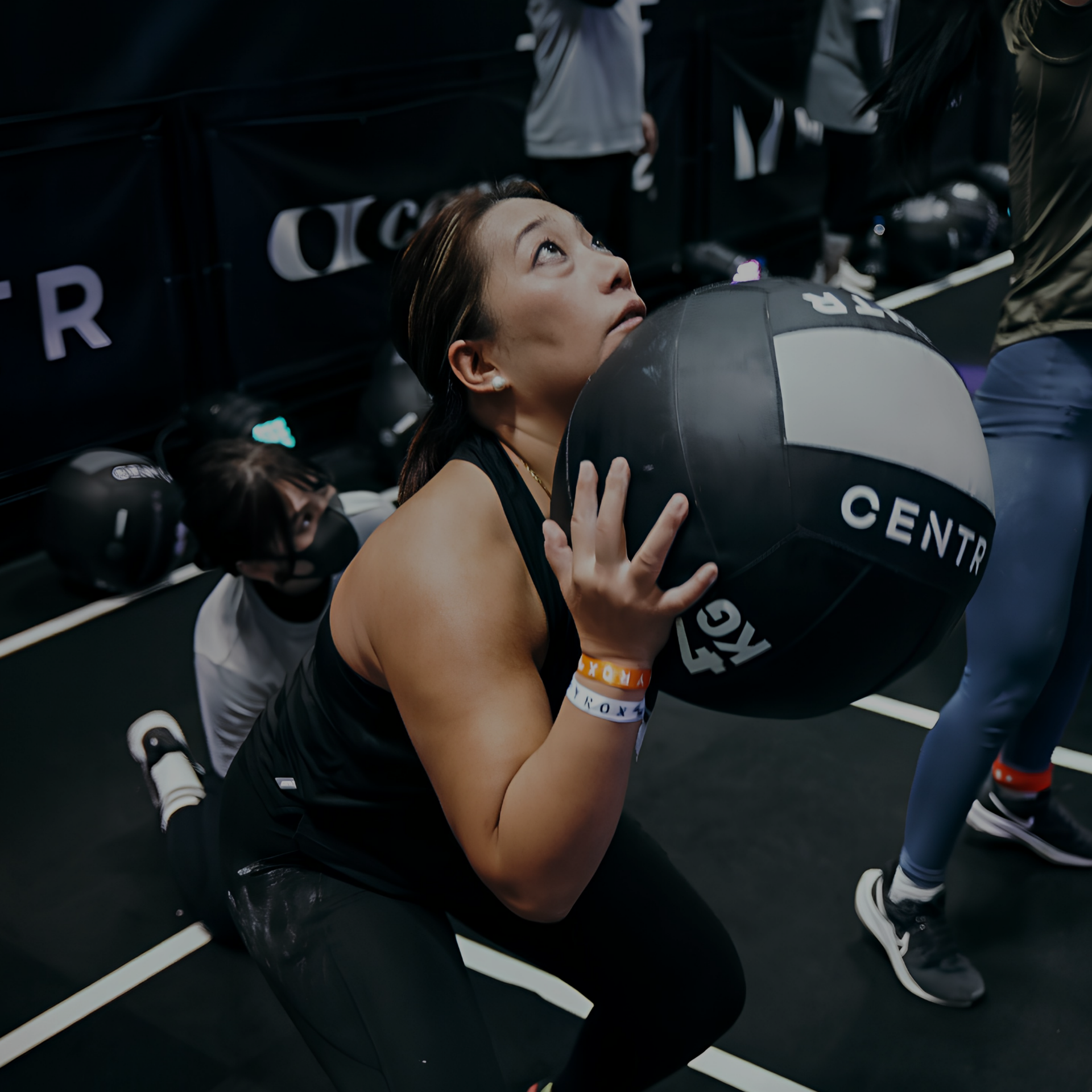 Woman lifting a black medicine ball in a gym, with another person behind her and workout gear nearby