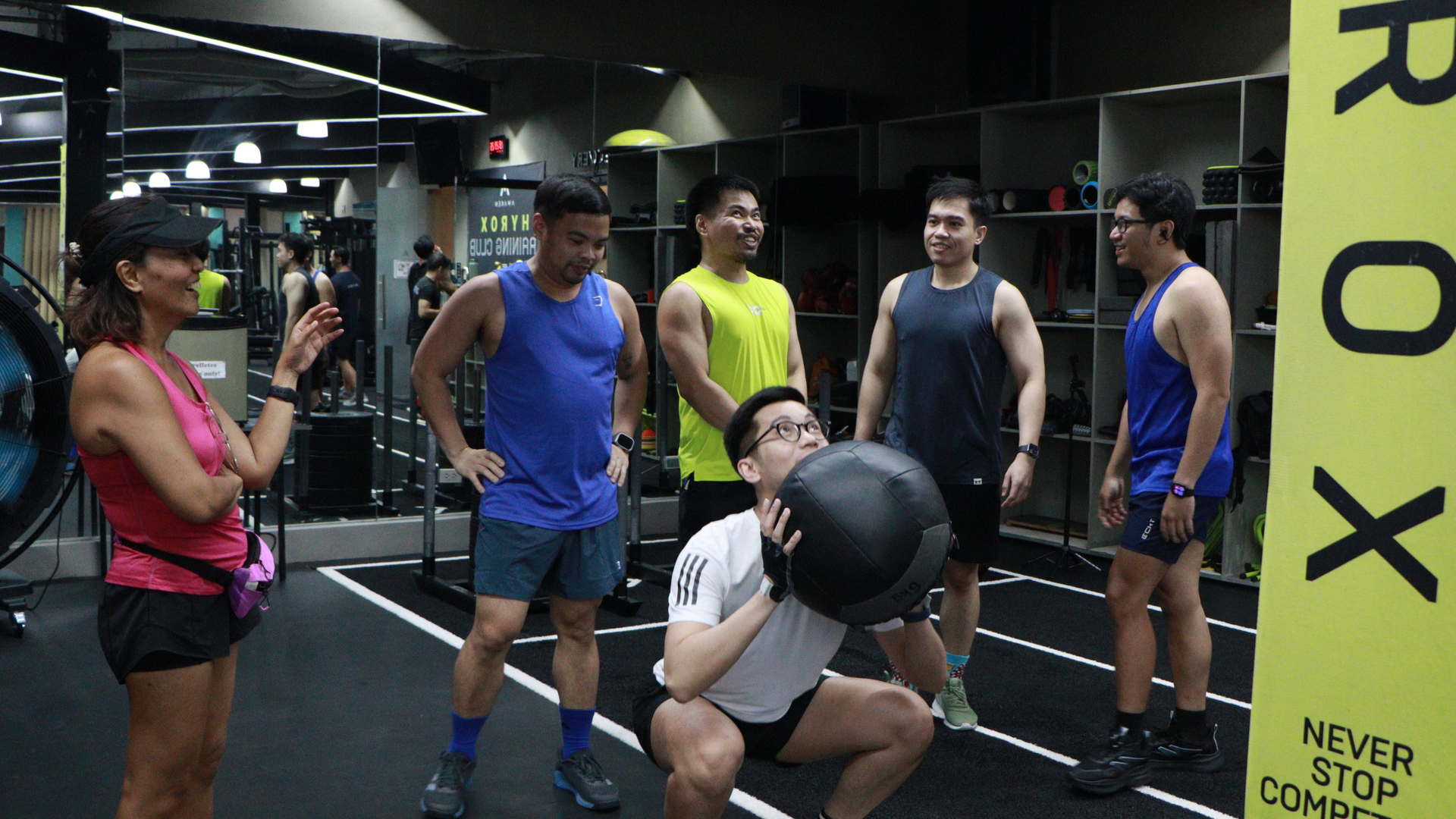 Gym class with people exercising, one person doing a squat while others watch near a yellow TRX strap.