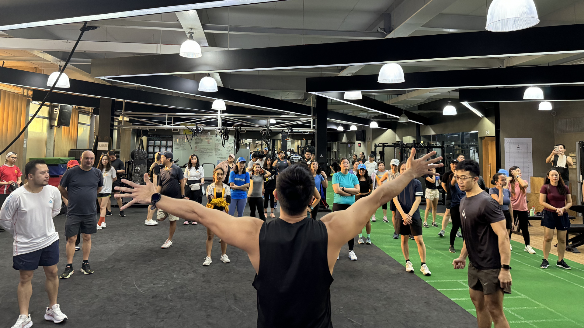 Group exercise class in a gym, instructor in foreground with arms raised and participants standing on mats.