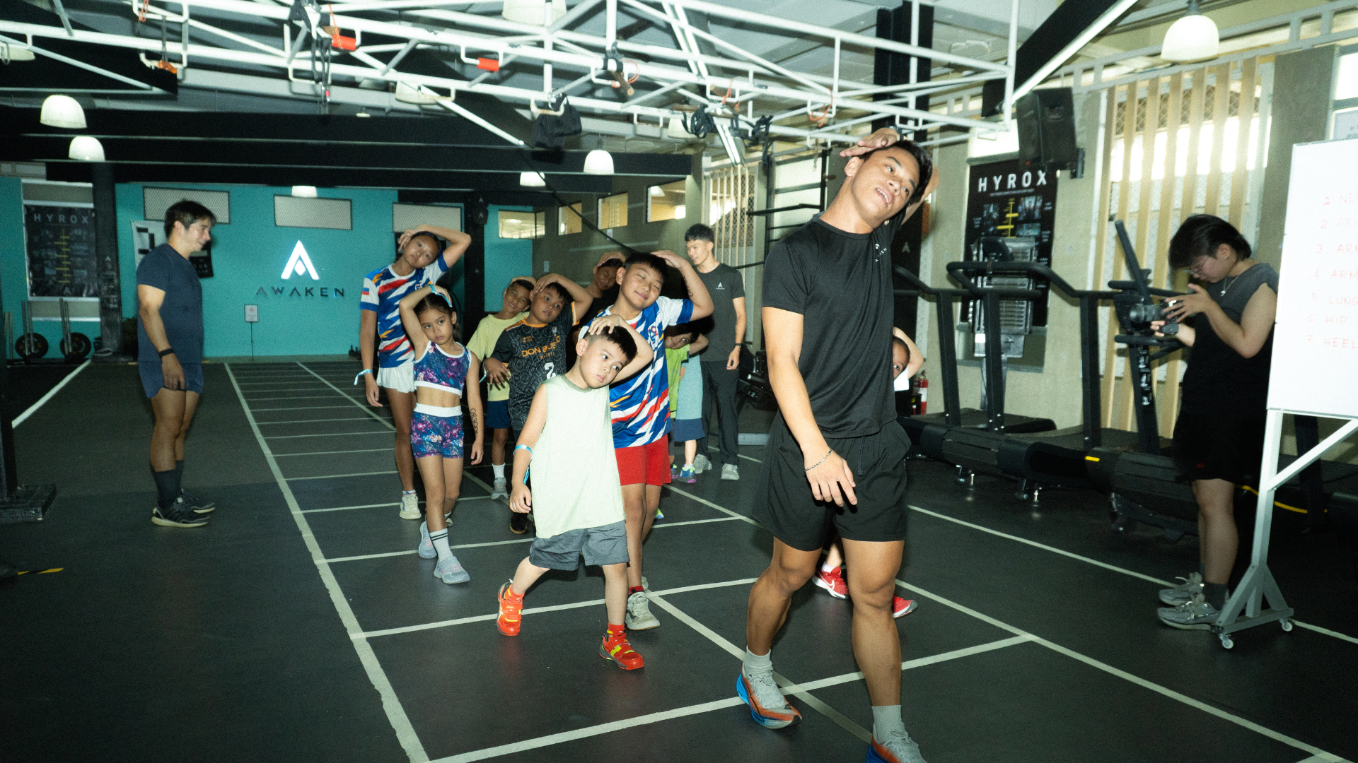 Group exercising indoors on marked floor lanes, led by a coach in black.