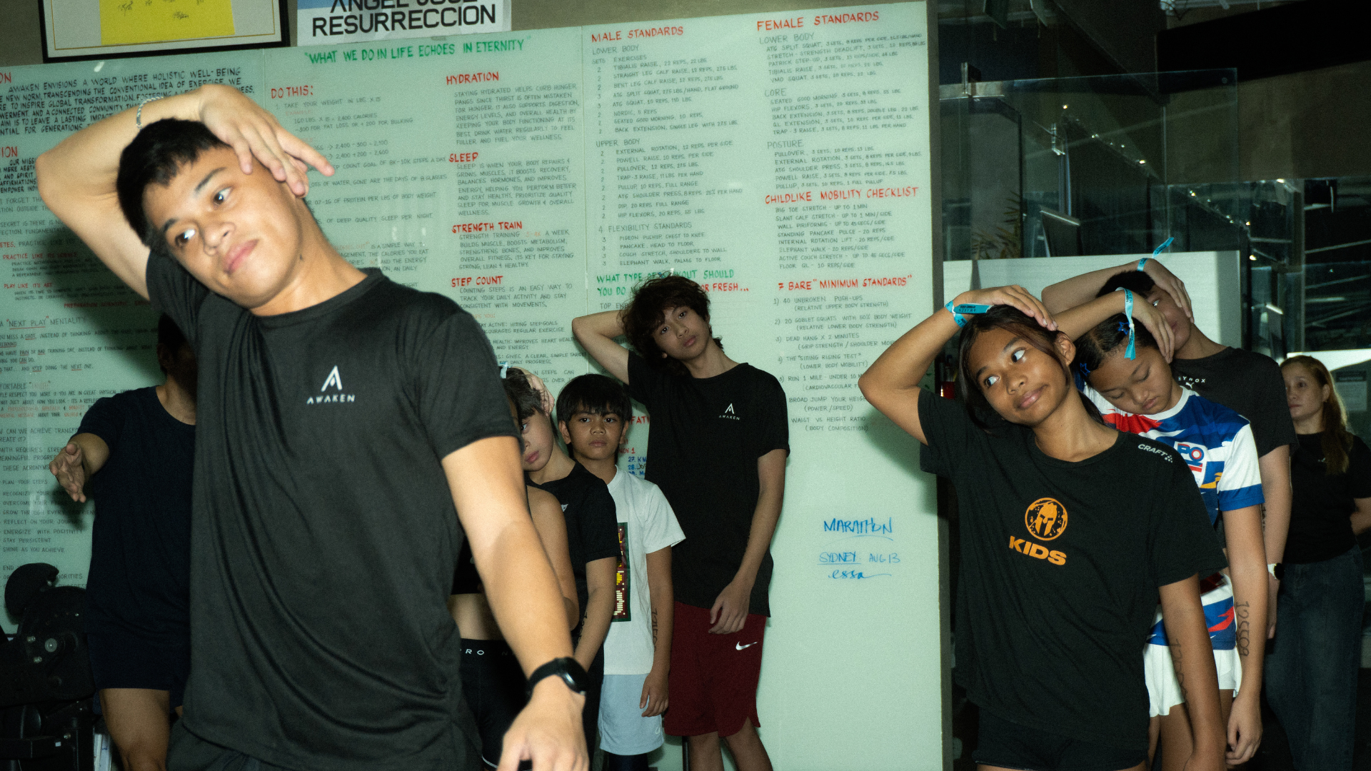 People in a gym class stretch their necks with arms overhead in front of a whiteboard.