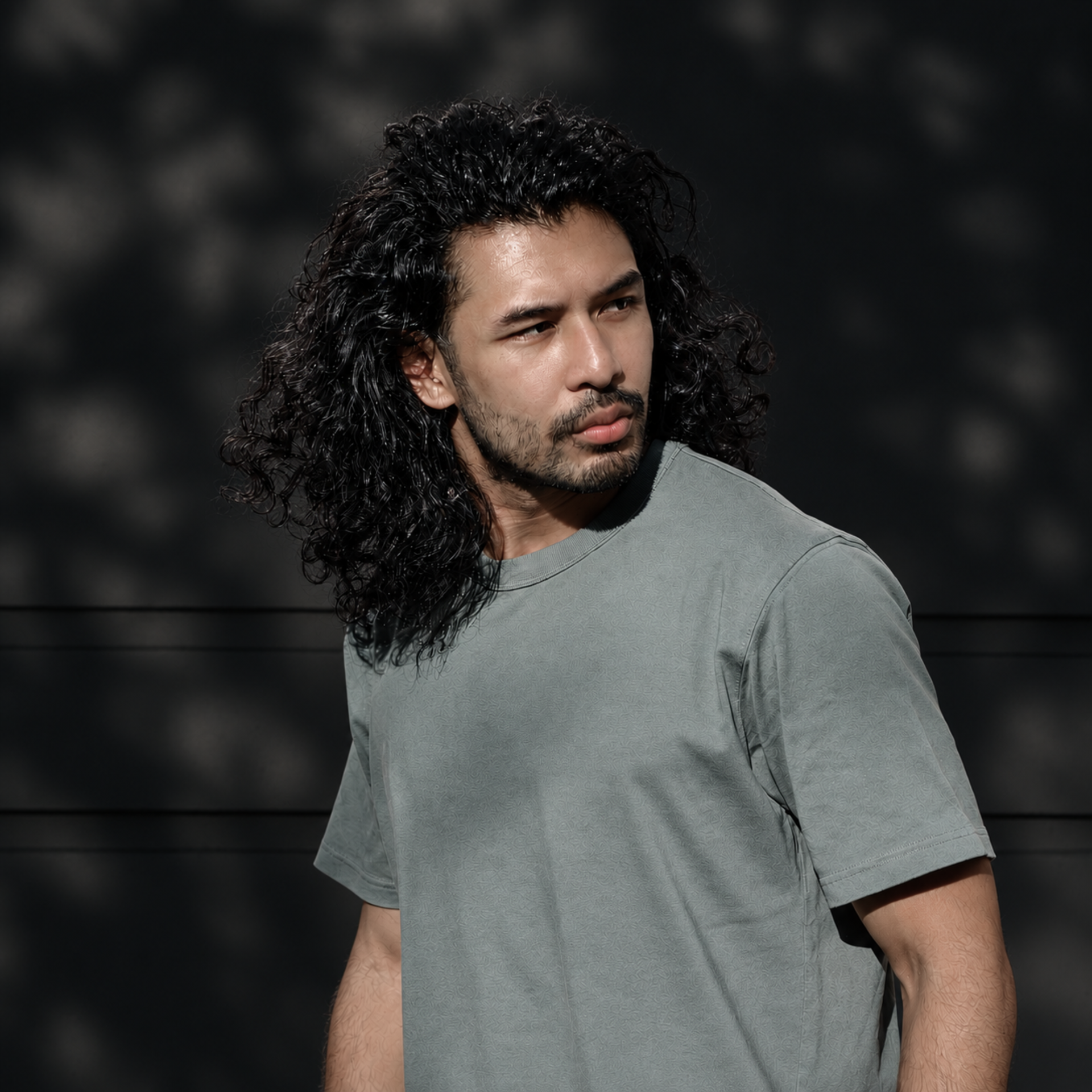 Man with long curly hair in a gray T-shirt, looking to the side against a dark background