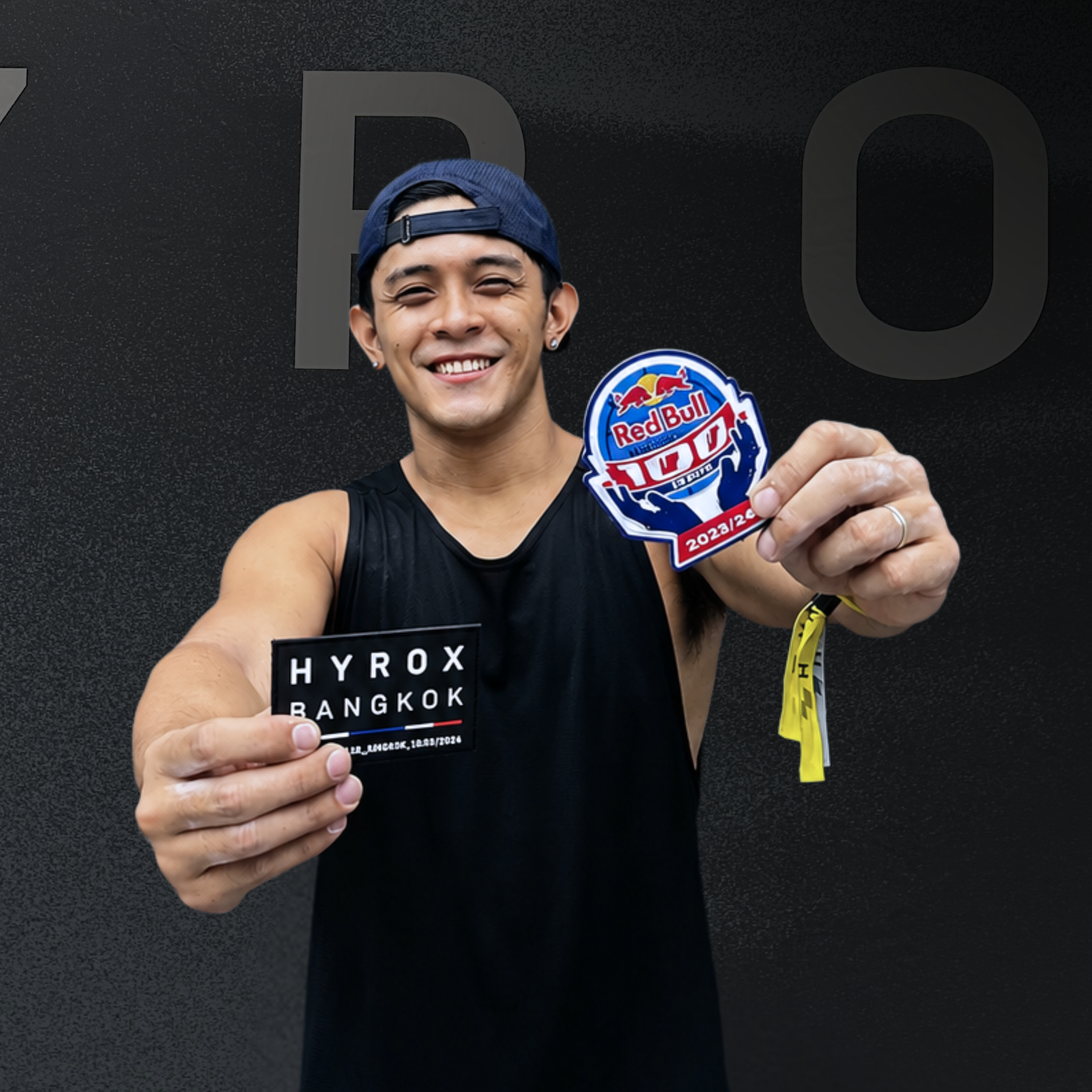 Smiling athlete in black HYROX Bangkok shirt holds two event patches toward the camera.